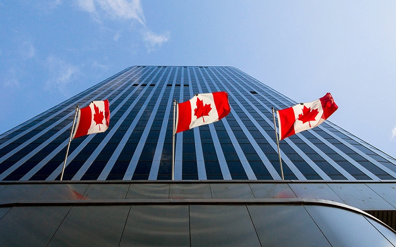 Three Canadian flags wave in front of a tall, modern glass skyscraper, viewed from below against a blue sky.