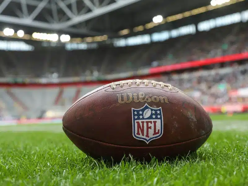 A Wilson NFL football rests on green grass in a stadium, with blurred crowds and stadium seating visible in the background.